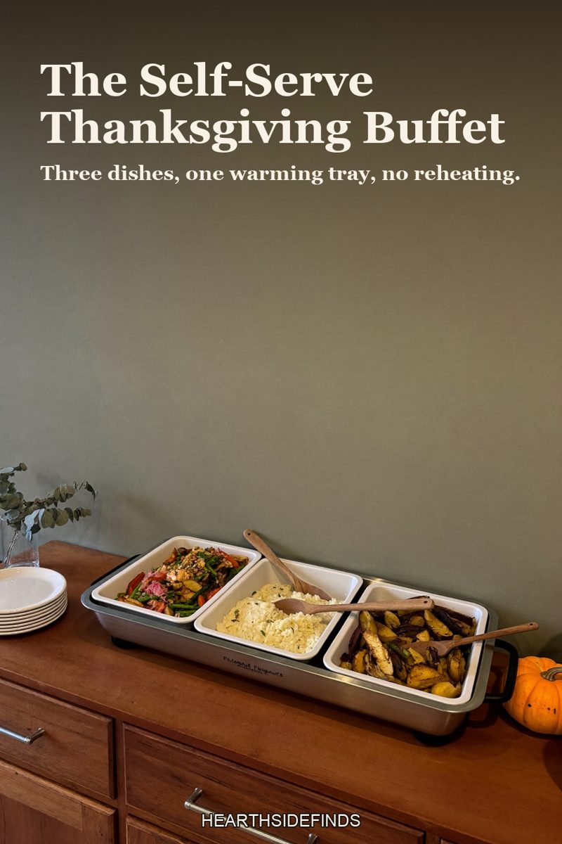 Wood sideboard with a stainless electric buffet server holding three dishes of food, a stack of white plates and a small pumpkin beside it, sage wall above.