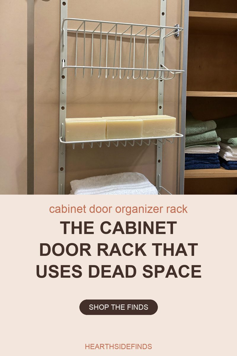 Photo of a white wire organizer rack hung on two vertical peg rails down the inside face of a tall cabinet door, an empty slotted wire basket on the upper tier, three plain cream soap blocks lying side by side on the tier below it, and the top of a stack of folded white towels in the basket at the bottom of the frame, with open wooden shelves holding folded green, white and navy towels beside the door on the right