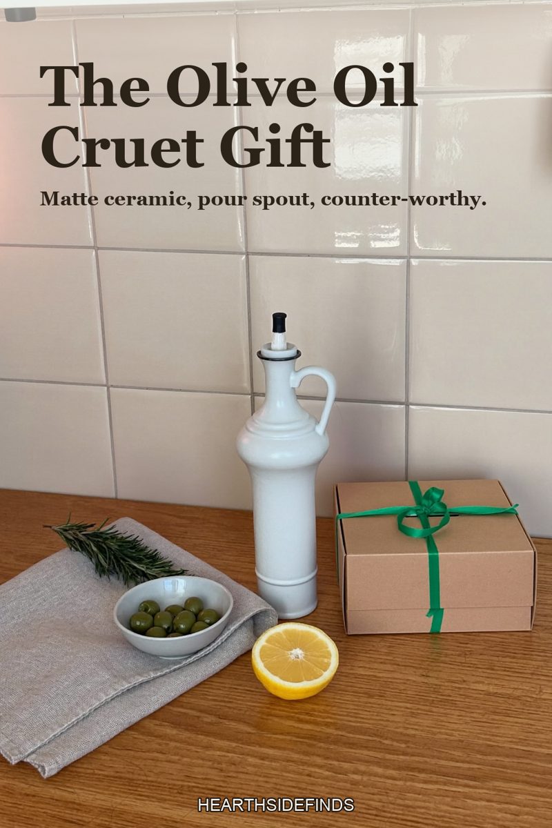 Matte white ceramic olive oil cruet with a pour spout on a wooden counter, a small dish of olives, a halved lemon, a linen cloth and a kraft gift box with green ribbon beside it.