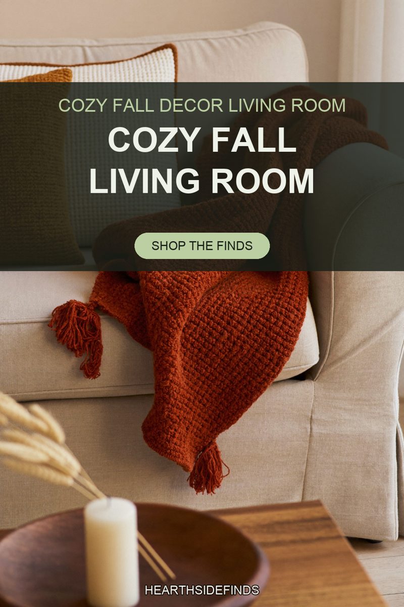 A chunky rust knit throw with corner tassels draped over the rolled arm of a cream linen sofa, one tassel resting on the seat cushion and the other hanging past the arm, two textured pillows in the sofa corner behind it, and a wooden bowl holding a pale pillar candle beside dried wheat stems on the coffee table in the foreground.