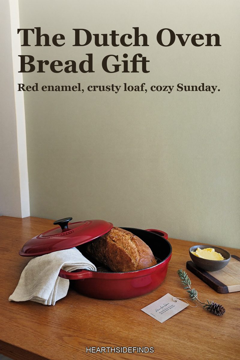 Red enameled cast iron dutch oven with a rustic loaf of bread inside on a wooden table, a linen towel, a wooden board with butter and a pine sprig beside it.