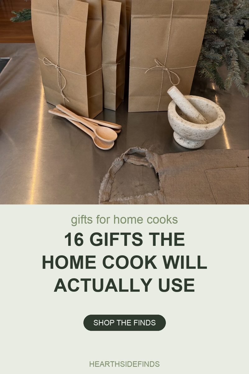 Photo of tall plain kraft paper bags wrapped in twine, two of them tied in bows, standing on a steel kitchen counter, a small stack of light wooden spoons lying beside them, a pale stone mortar with its pestle to the right, a loose piece of frayed natural linen across the foreground and a blue spruce branch at the right edge of the frame