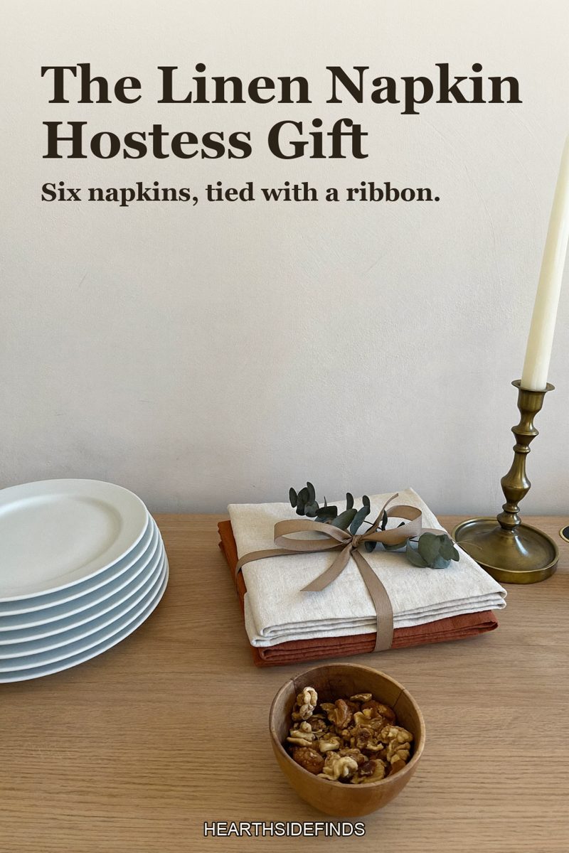Folded natural linen napkins in cream and rust tied with a ribbon and eucalyptus on a light wood table, a stack of white plates, a brass candlestick and a bowl of walnuts beside them.