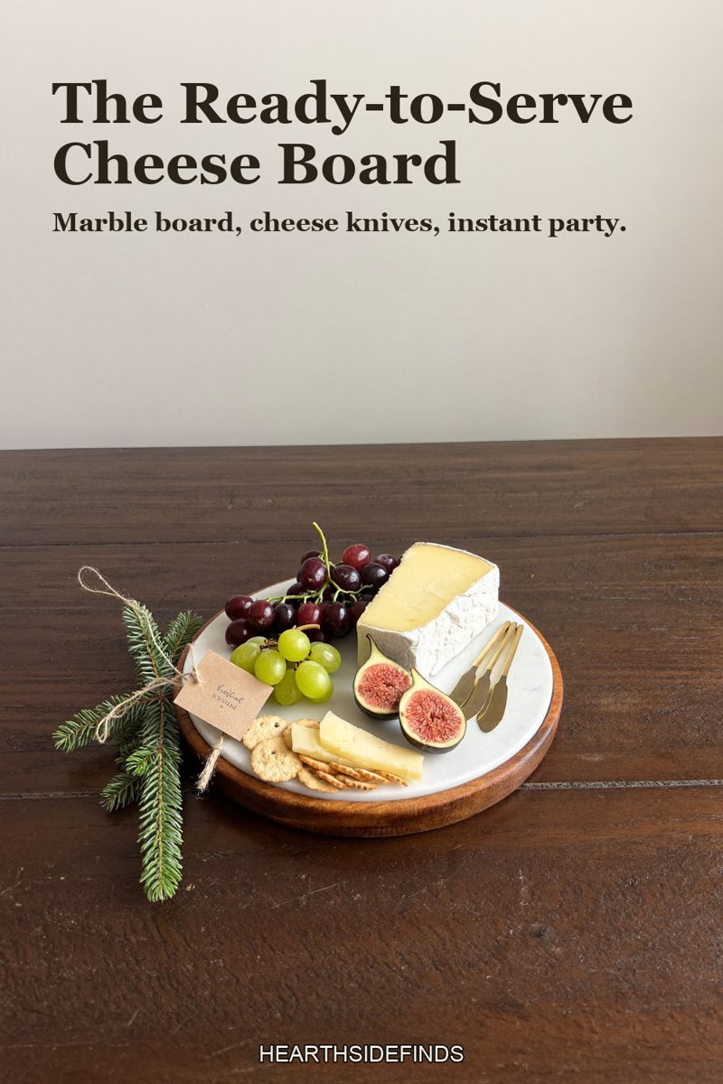 Round marble cheese board with a wood edge holding cheeses, grapes, crackers and figs with small brass knives on a dark wood table, a fir sprig beside it.