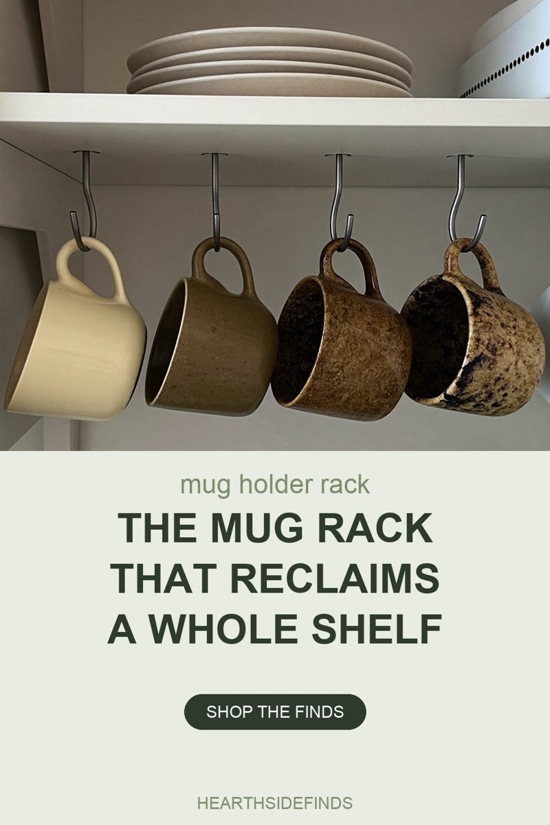 Photo of four ceramic mugs hanging by their handles from steel S-hooks screwed into the underside of a white kitchen shelf — one pale cream, one olive brown, one mottled tan and one speckled black and cream — with a stack of four pale stoneware plates and the edge of a white perforated canister on the shelf above them