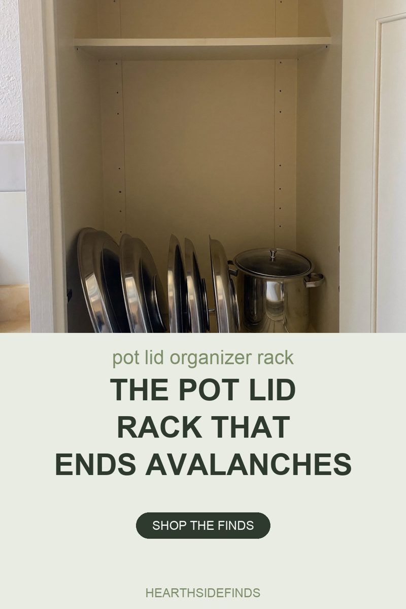 Photo into an open cream kitchen cabinet where a row of stainless steel pot lids stands on edge in a low wire rack resting on the cabinet floor, a lidded stainless stockpot stands beside them on the right, and the shelf directly above them is empty