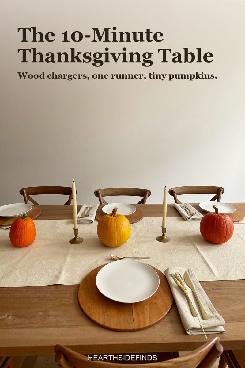 Wooden dining table with round wood charger plates under white plates, a cream linen runner, mini orange pumpkins and cream taper candles in brass holders.