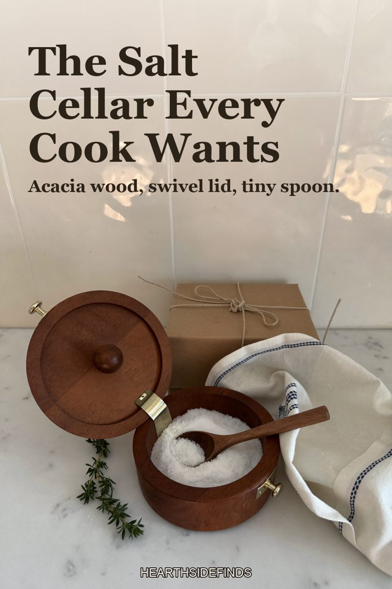 Open acacia wood salt cellar with a swivel lid and a small wooden spoon in coarse salt on a marble counter, a linen cloth, thyme and a kraft gift box behind it.