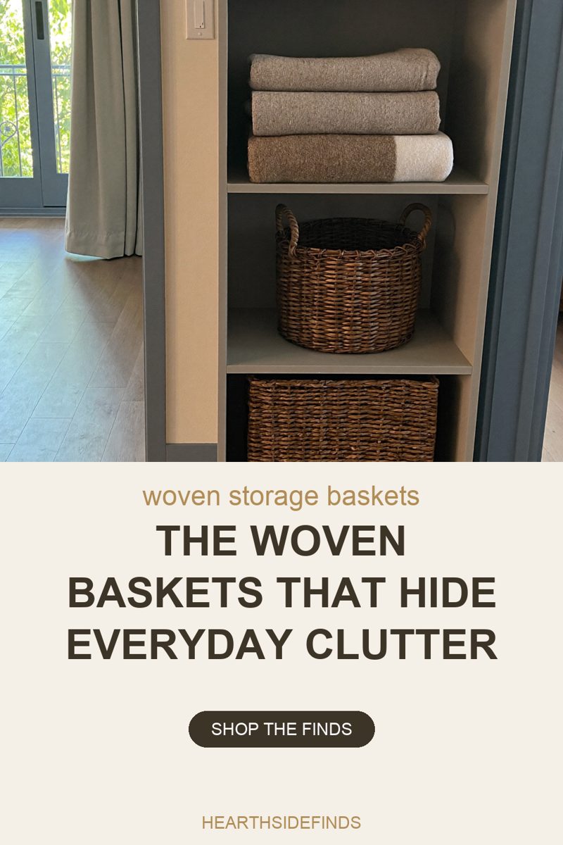 Photo of a putty-grey open shelf unit standing against a cream wall beside a curtained glass balcony door, pale wood flooring filling the left of the frame: the top cubby holds three folded waffle towels in grey, taupe and cream, the middle cubby a round woven wicker basket with two rope handles, and the bottom cubby a deeper rectangular seagrass basket filling it wall to wall with its lower half cut off by the bottom edge of the frame, a blank pale light switch plate on the wall at the top left corner of the unit