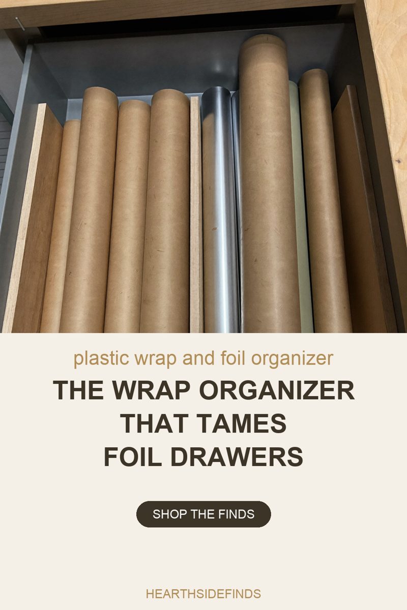 Looking down into an open kitchen drawer lined in grey metal, with the drawer's back wall and its left wall in frame and a wooden cabinet rail running across the top. Eight rolls lie flat inside it, running away from the camera with their round cut ends toward the back: six plain brown kraft, one silver foil roll at the centre, and one narrow pale sage roll to the right of it. Boards stand on edge as dividers, one pale plywood between the kraft rolls and the foil roll, one pale plywood closing the left side and one dark brown closing the right. A pale plywood cabinet panel fills the right edge. Nothing in the frame carries lettering or a label.